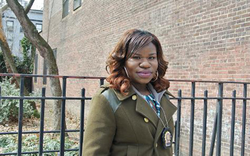 Vanessa Okonta in a green coat in front of a brick building and gate. There is a badge hanging from a chain around her neck.